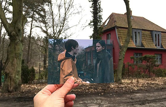 Eine Hand hält ein ausgedrucktes Foto von der Serie Dark vor ein rotes Haus in einem Wald.