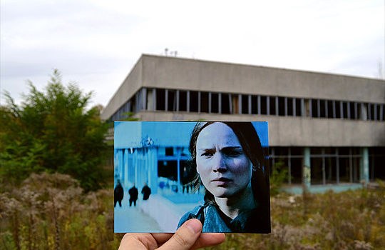 Eine Hand hält ein ausgedrucktes Foto aus dem Film "Tribute von Panem" vor einer verlassenen Fabrikhalle.