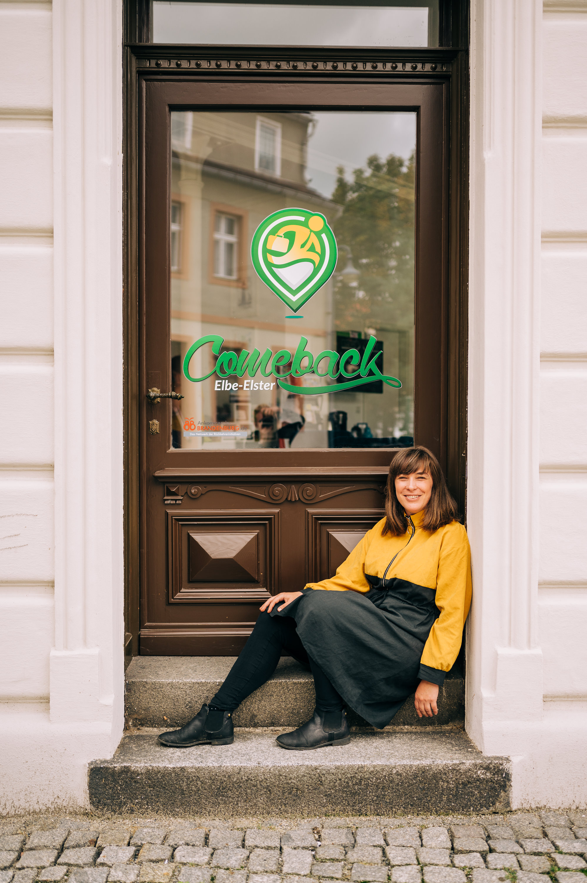 Eine junge Frau sitzt lächelnd in einem Hauseingang. Auf der Eingangstür steht bedruckt "Comeback Elbe Elster".