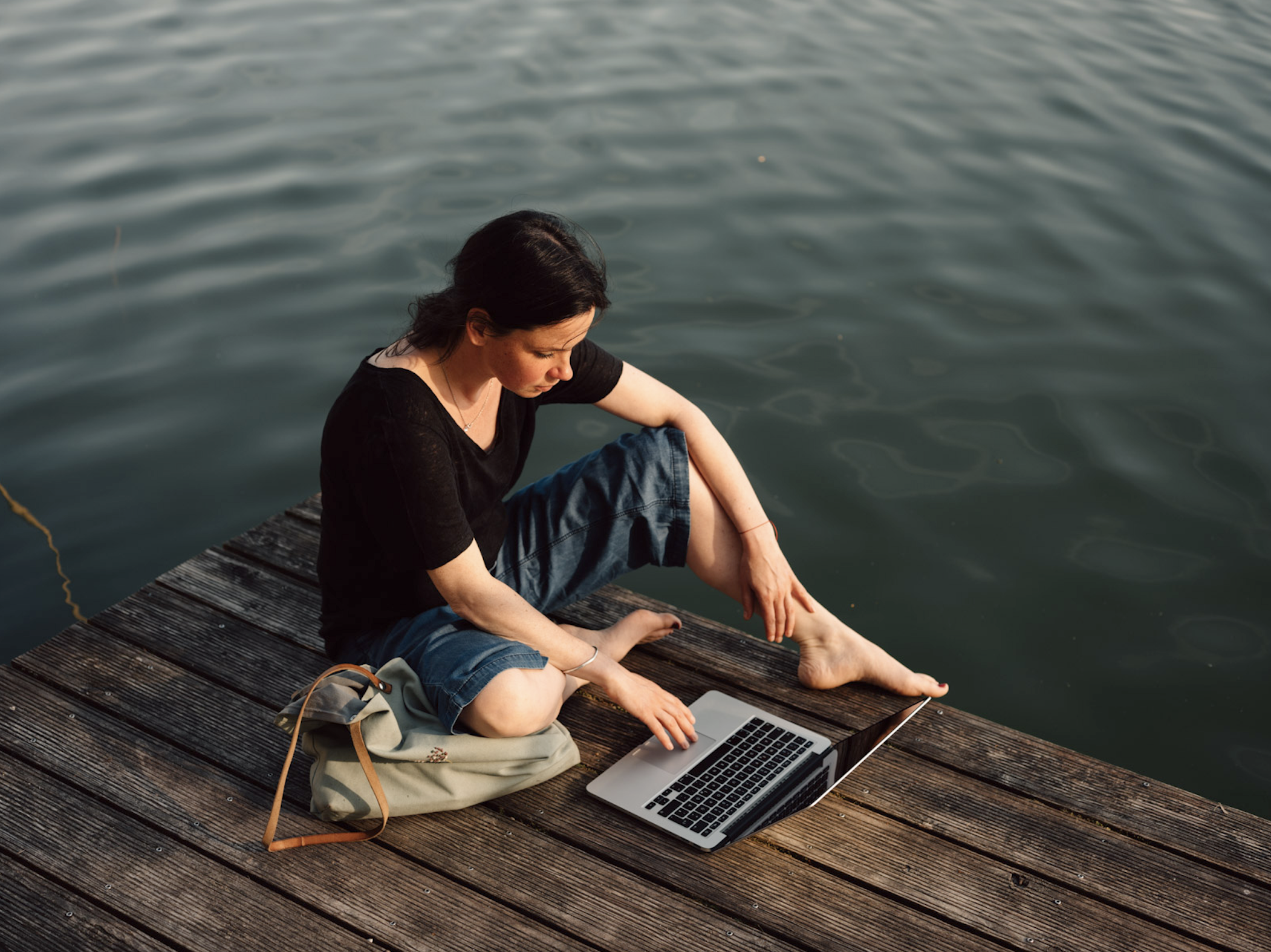 Eine Frau sitzt mit ihrem Laptop auf einem Steg am Wasser.