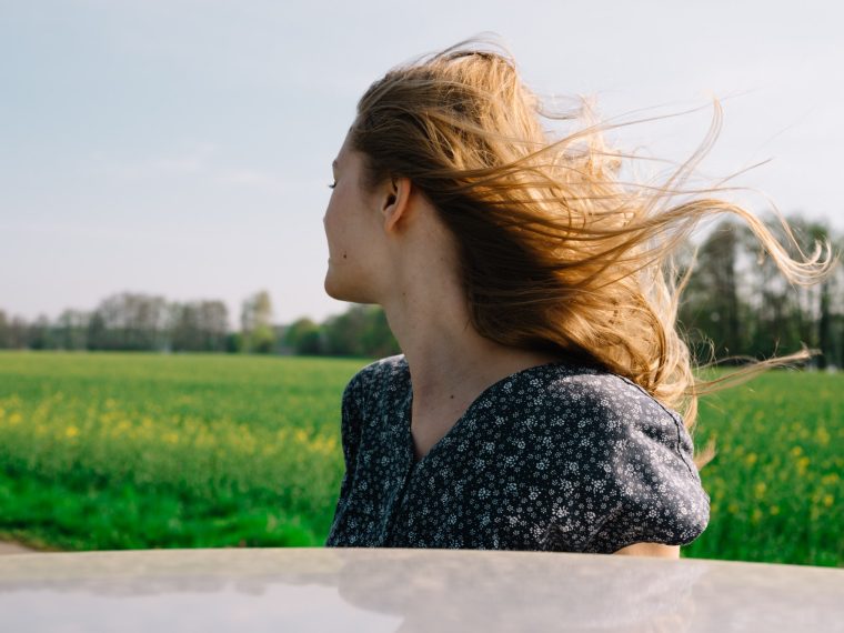 Eine Frau blickt über ein grünes Feld. Ihre Haare werden vom Wind verweht.