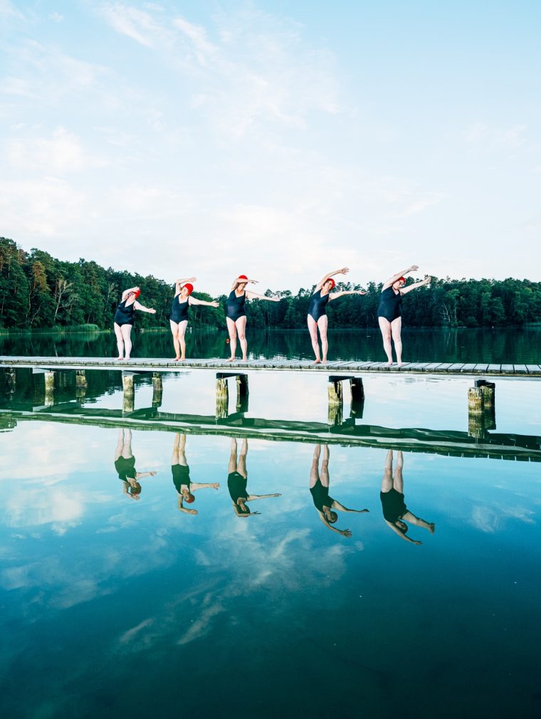 Fünf Damen in schwarzen Badeanzügen und roten Schwimmkappen machen auf einem Steg auf einem See einheitliche Schwimmbewegungen.