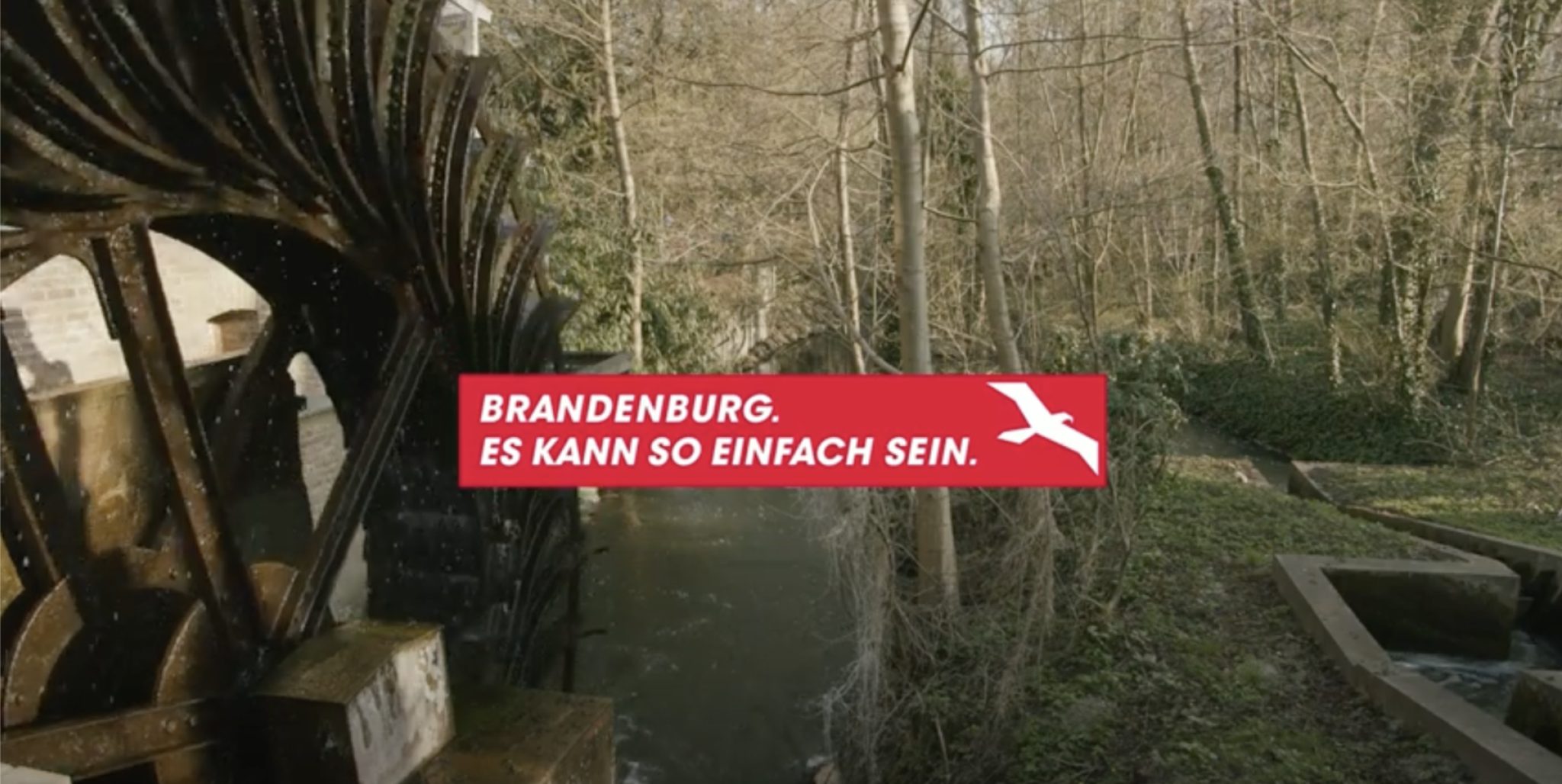 Ein Mülenrad betrieben von einem kleinen Bach. Im Hintergrund ein Wald mit Bäumen ohne Laub. Eine Aufschrift "Brandenburg. Es kann so einfach sein."