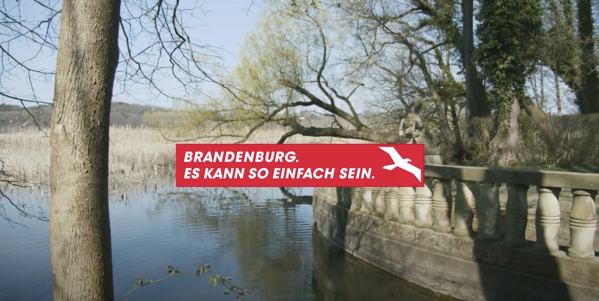 Ein See mit viel Schilf, links im Eck steht ein Baum. Auf der rechten Seite ist ein Geländer aus Stein und Bäume im Hintergrund. Im Vordergrund die Aufschrift "Brandenburg. Es kann so einfach sein."