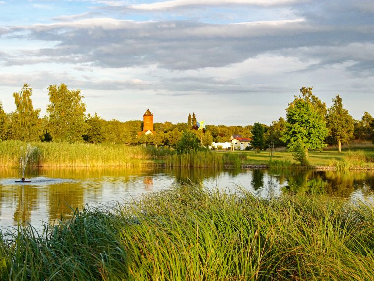 Ein kleiner See im Abendlicht mit viel Gras drumherum. Im Hintergrund ist ein Dorf mit Kirche.