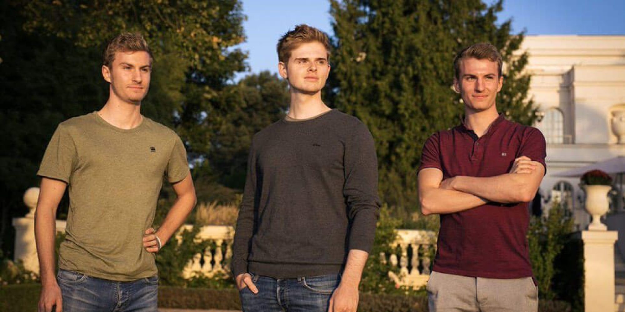 Drei junge Männer stehen vor Bäumen und einem herrschaftlichen Haus.