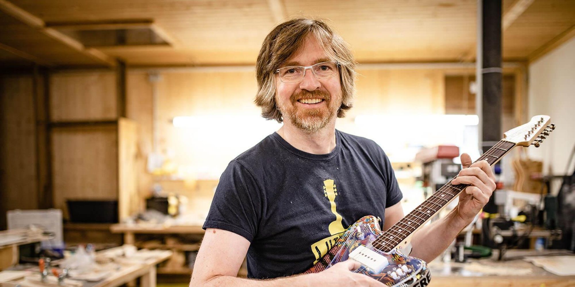 Ein Mann steht in einer Werkstatt und schaut lächelnd in die Kamera. In der Hand hält er eine bunte E-Gitarre.