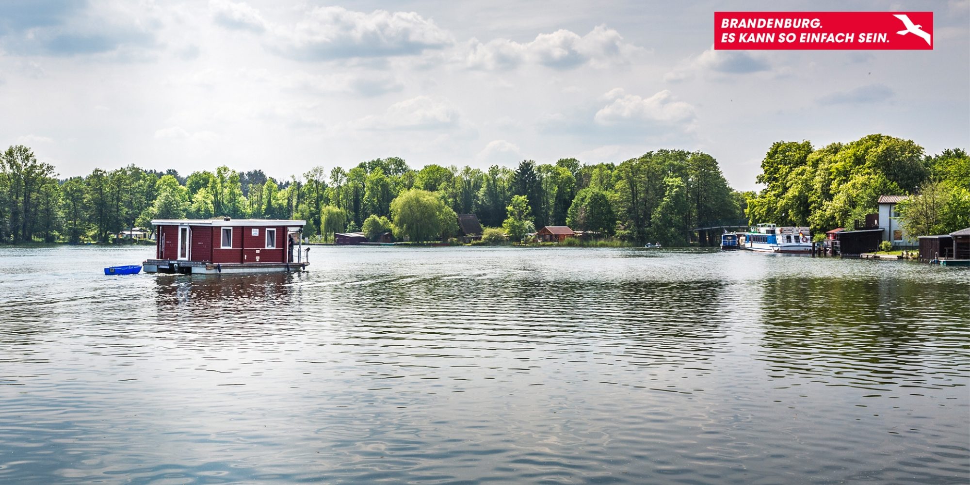 Ein ruhiger See an einem hellen Tag. Auf dem See fährt ein kleines Hausboot. Im Hintergrund Wald.