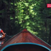 Eine Kanu-Spitze auf einem schmalen Fluss zwischen Bäumen. Im Hintergrund zwei Menschen in einem Kayak.