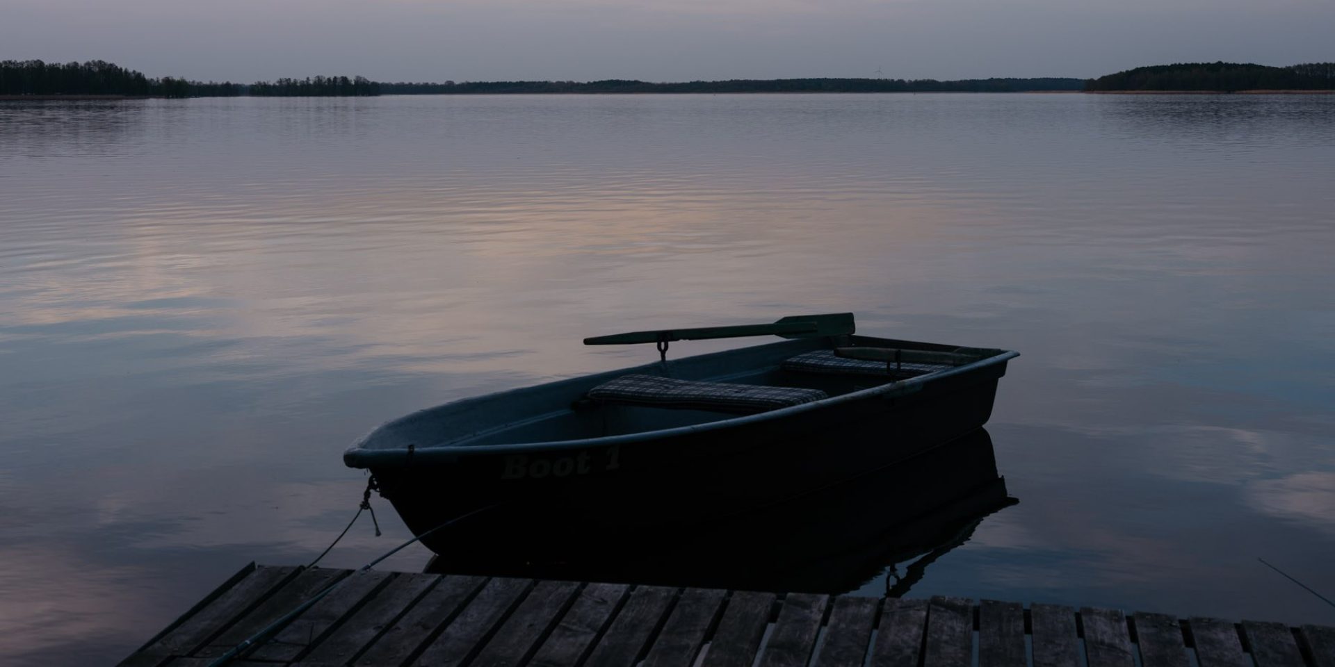 Ein ruhiger See am Abend. Darauf ein leeres Ruderboot, das an einem Steg befestigt ist.