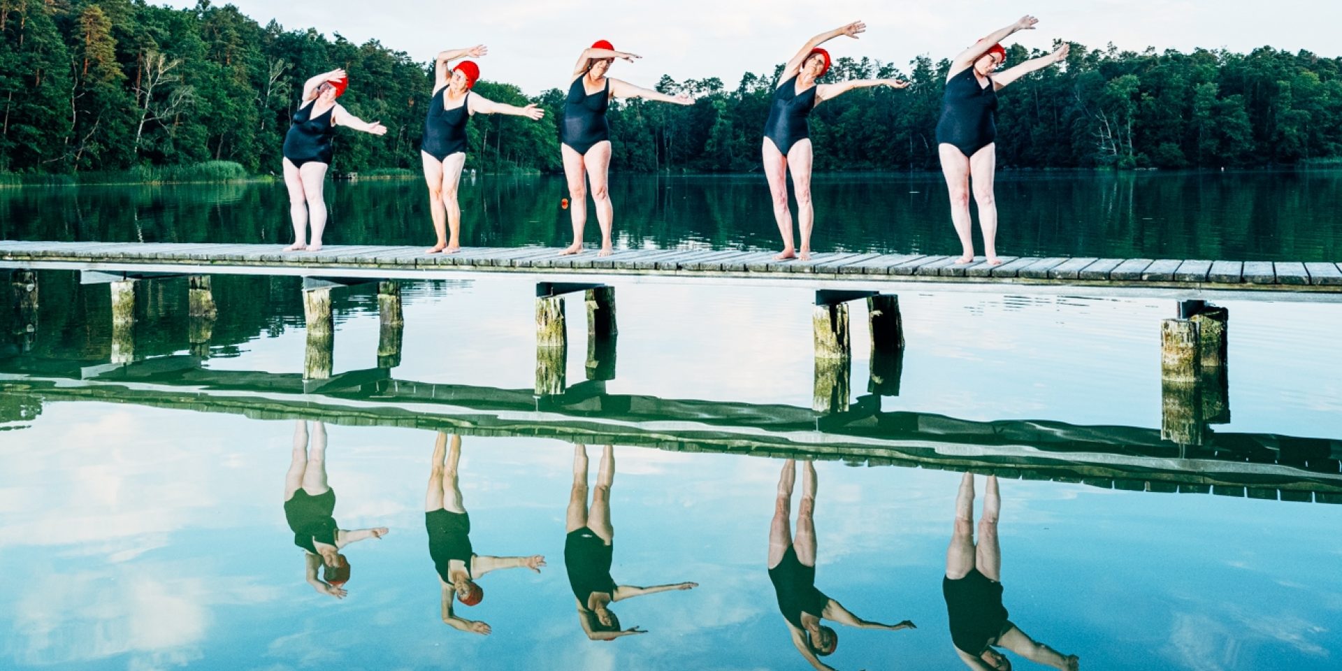 Fünf Damen in schwarzen Badeanzügen und roten Schwimmkappen machen auf einem Steg auf einem See einheitliche Schwimmbewegungen.