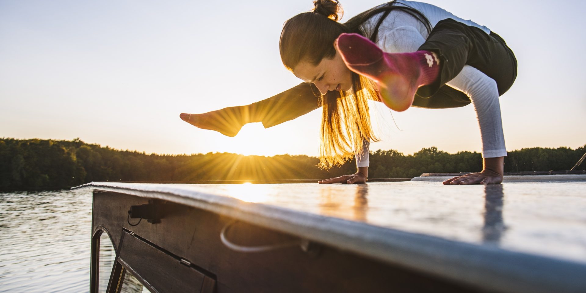 Eine Frau macht auf einem Boot eine Yoga-Pose. Im Hintergrund sieht man einen See und einen Wald.