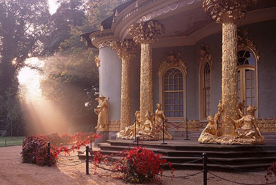 Ein schöner barocker Schlosseingang in einem Park mit goldenen Säulen und Statuen.