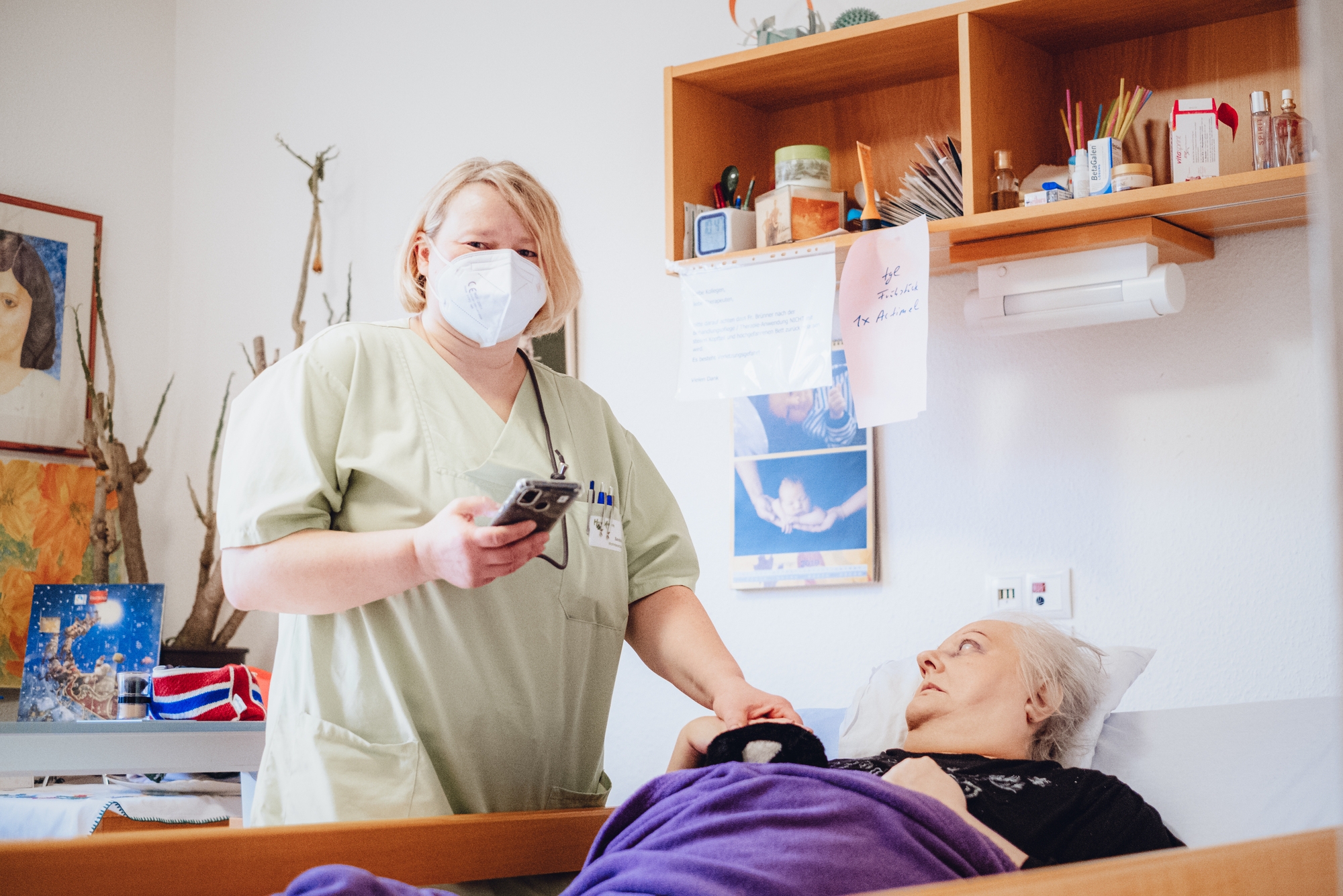 Eine Pflegekraft steht mit Handy in ihrer Hand bei einer Patientin am Bett.